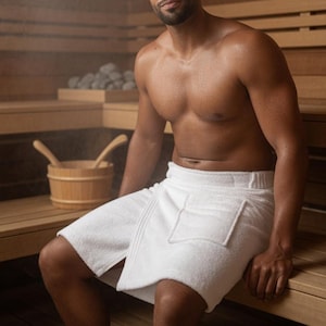 May include: A person wearing a white spa towel with a pocket, sitting in a wooden sauna. A wooden bucket with two wooden ladles is visible. The sauna has wooden benches and walls. Steam is visible in the background.