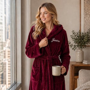 May include: A burgundy hooded robe with a matching belt and pockets. The robe has the name "Jessica" embroidered on the chest. The plush robe is knee-length and is made of a soft, velvety material. A mug is held in the hand.
