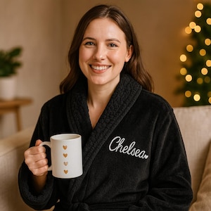 May include: A woman in a black plush robe embroidered with the name "Chelsea" in white, holding a white mug with four gold hearts. A Christmas tree with lights is visible in the background.