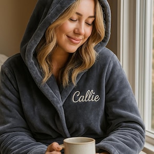 May include: A dark gray hooded robe with the name "Callie" embroidered in white. The robe is made of a soft, plush material. A person is holding a beige mug.