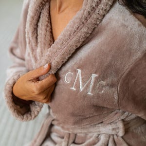 May include: A plush, mauve-colored robe with a shawl collar and a matching belt. The robe features white embroidered initials on the chest. The soft, textured fabric suggests comfort and relaxation, perfect for lounging.