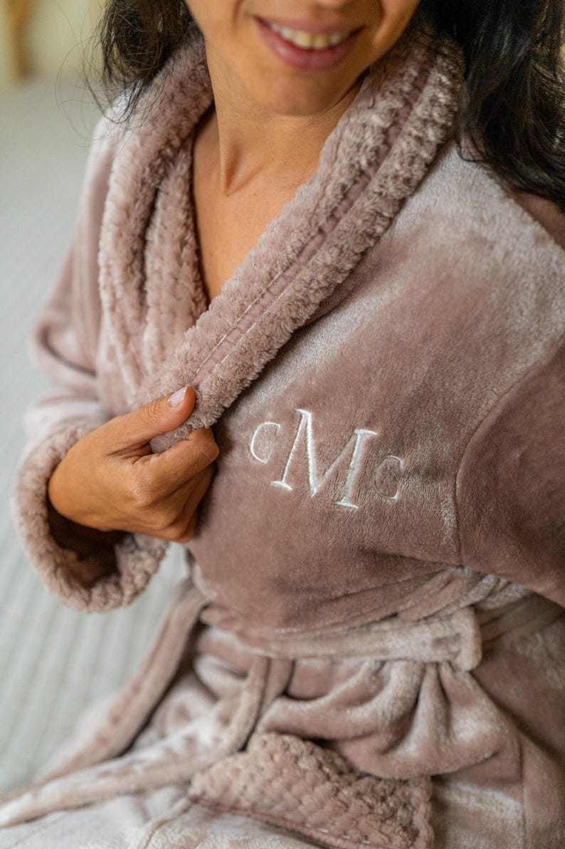 May include: A plush, mauve-colored robe with a shawl collar and a matching belt. The robe features white embroidered initials on the chest. The soft, textured fabric suggests comfort and relaxation, perfect for lounging.