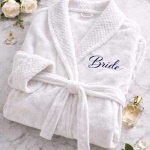 May include: A white plush robe with the word "Bride" embroidered in navy blue. The robe is tied with a matching belt. The image includes white roses, perfume, and jewelry, suggesting a bridal theme.