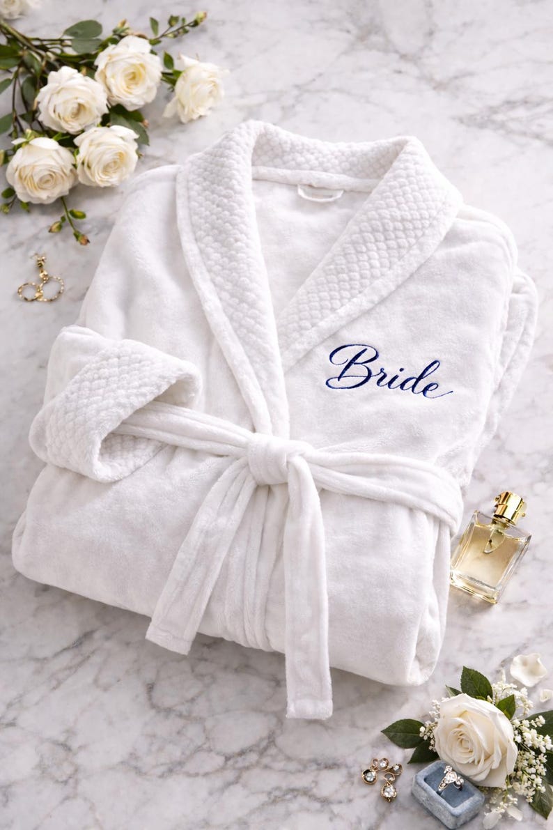 May include: A white plush robe with the word "Bride" embroidered in navy blue. The robe is tied with a matching belt. The image includes white roses, perfume, and jewelry, suggesting a bridal theme.