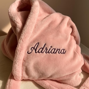 May include: A light pink plush robe with the name "Adriana" embroidered in a darker pink script. The robe is folded, showing the soft texture of the fabric and the shawl collar. The overall aesthetic is cozy and luxurious.