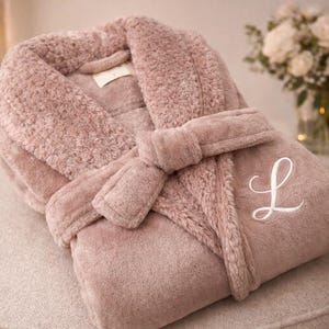 May include: A plush, dusty rose-colored robe with a matching belt, folded neatly on a neutral-toned surface. The robe features a white embroidered letter "L" on the front. The soft, textured fabric suggests comfort and relaxation.