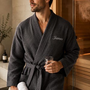 May include: Charcoal gray waffle-weave robe with the name "James" embroidered on the chest. The robe has a matching belt and pockets. A rolled white towel and a glass of water are held in the hands.