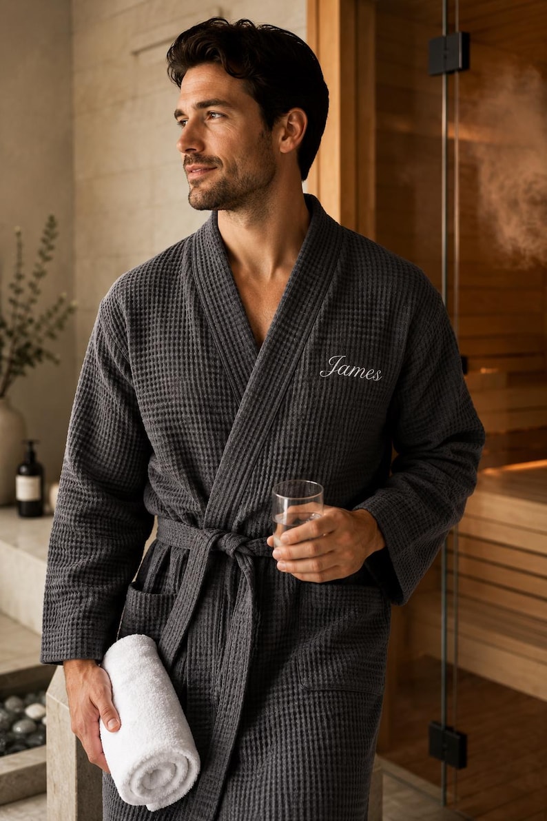 May include: Charcoal gray waffle-weave robe with the name "James" embroidered on the chest. The robe has a matching belt and pockets. A rolled white towel and a glass of water are held in the hands.