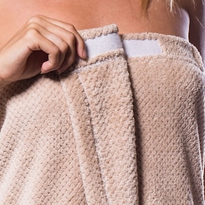 May include: A beige plush towel with two white straps. The towel is wrapped around a person's waist.