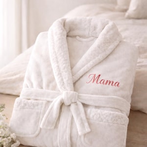May include: A white plush robe with a shawl collar and a tie belt. The word "Mama" is embroidered in pink on the chest. The robe is folded neatly on a wooden surface.