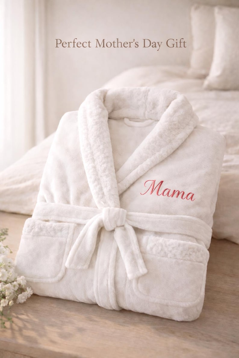May include: A white plush robe with a shawl collar and a tie belt. The word "Mama" is embroidered in pink on the chest. The robe is folded neatly on a wooden surface.