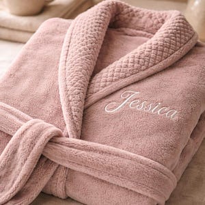May include: A dusty rose-colored plush bathrobe with a shawl collar and a matching belt. The word "Jessica" is embroidered in white script on the front. The robe is folded neatly, suggesting a spa or relaxation theme.