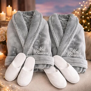 May include: Two gray plush bathrobes and matching white slippers are displayed. Each robe is embroidered with "Mrs. Dawson" in elegant script. The robes have a textured collar and cuffs. The slippers are soft and fluffy, suggesting comfort and relaxation.