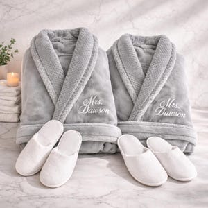 May include: Two gray plush bathrobes with the embroidered name "Mrs. Dawson" and two pairs of white slippers. The robes have shawl collars and are displayed on a marble surface. A candle and plant are in the background.