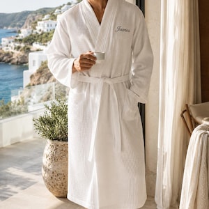 May include: White waffle-weave robe with the name "James" embroidered on the chest. The robe has a shawl collar, a waist tie, and pockets. The person is holding a white cup. The robe is made of a textured fabric.