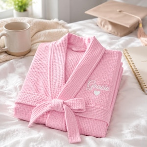 May include: A pink waffle-weave dressing gown with a matching tie belt. The gown is embroidered with the name "Gracie" in white script above a small white heart. The gown is folded neatly on a white bedspread.