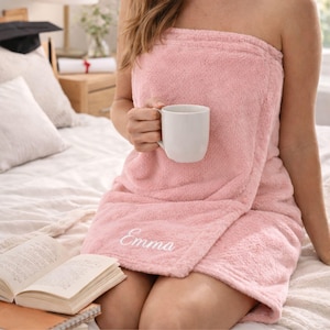 May include: A light pink, plush spa towel wrap with the name "Emma" embroidered on the front. A white mug is held in the hand. A book and a graduation cap are in the background.
