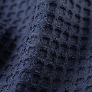 May include: Close-up of a navy blue waffle-weave fabric. The textured material has a repeating pattern of small, raised squares, creating a grid-like appearance. The fabric is soft and appears to be slightly draped, with subtle shadows adding depth.