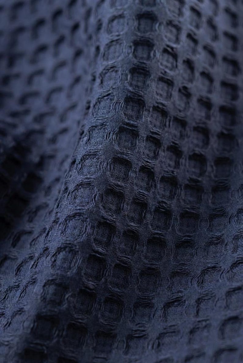 May include: Close-up of a navy blue waffle-weave fabric. The textured material has a repeating pattern of small, raised squares, creating a grid-like appearance. The fabric is soft and appears to be slightly draped, with subtle shadows adding depth.