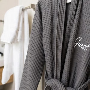 May include: A dark gray waffle-weave bathrobe with the word "Guest" embroidered in white. The robe is hanging on a white hanger. A white towel is hanging on a towel rack in the background.