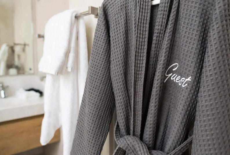 May include: A dark gray waffle-weave bathrobe with the word "Guest" embroidered in white. The robe is hanging on a white hanger. A white towel is hanging on a towel rack in the background.