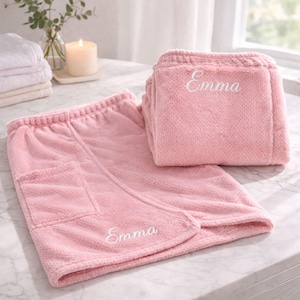 May include: A set of two pink terry cloth spa wraps, one folded and one open. The open wrap has a pocket and the name "Emma" embroidered in white. The folded wrap also has the name "Emma" embroidered on it. The wraps are on a marble surface.