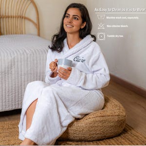 May include: A white bathrobe with the name "Claire" embroidered on the chest. The robe is worn by a person sitting on a woven pouf, holding a patterned mug. The image includes text: "As Easy to Clean as it is to Wear".