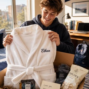 May include: A white plush bathrobe with the name Ethan embroidered in black script on the chest, packaged in a gift box with a bag of sea salt popcorn, a pair of dark gray socks, a packet of Kuju coffee, and a Built protein bar with a note.