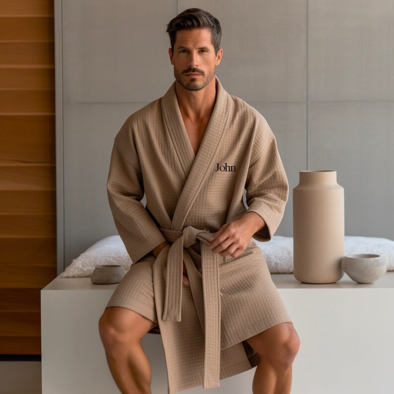 May include: A tan waffle-weave robe with a matching belt, personalized with the name "John" embroidered on the chest. The robe is knee-length and has wide sleeves. The person is sitting on a white surface.