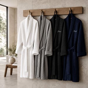 May include: Four waffle-weave bathrobes in white, gray, black, and navy blue hang on a wooden rack. The black and navy robes have embroidered names. Each robe has a matching belt. The background is a neutral-toned bathroom.