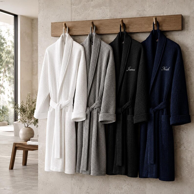 May include: Four waffle-weave bathrobes in white, gray, black, and navy blue hang on a wooden rack. The black and navy robes have embroidered names. Each robe has a matching belt. The background is a neutral-toned bathroom.