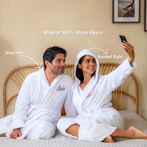 May include: Two white fleece bathrobes, one with a shawl collar and the other with a hood. The robes have embroidered names and are made of 100% warm fleece. One person is taking a selfie with a smartphone.