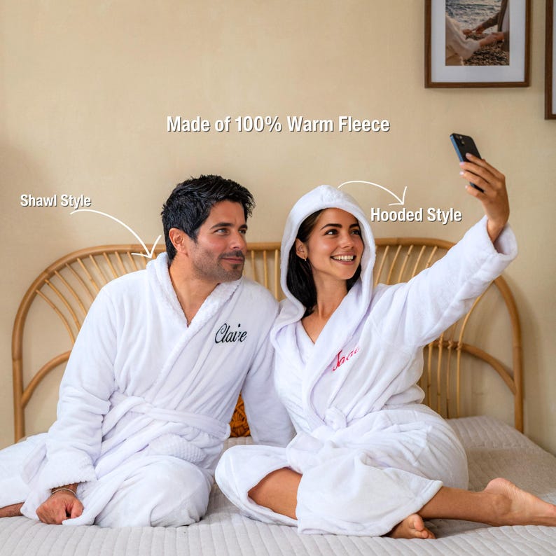 May include: Two white fleece bathrobes, one with a shawl collar and the other with a hood. The robes have embroidered names and are made of 100% warm fleece. One person is taking a selfie with a smartphone.