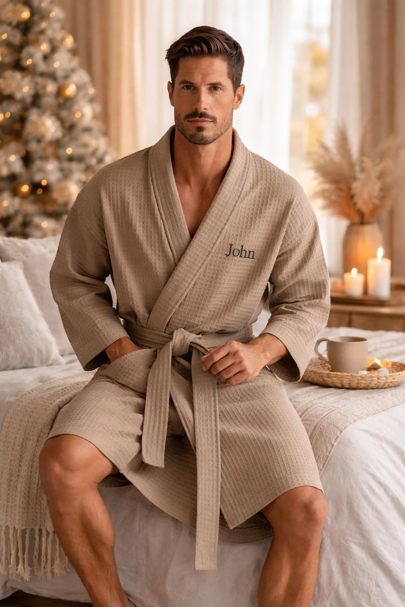 May include: A beige waffle-weave robe with a matching belt and pockets. The word "John" is embroidered on the chest. The robe is knee-length and worn open, with the model seated on a bed. The background includes a Christmas tree.