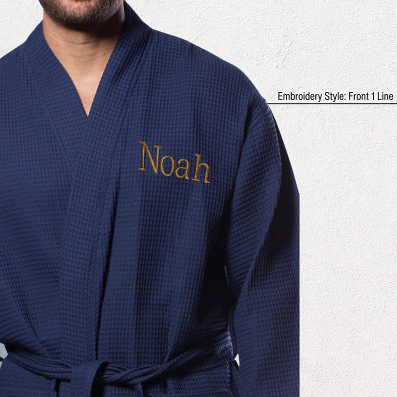 May include: Navy blue waffle-weave robe with the name "Noah" embroidered in gold thread on the chest. The robe has a shawl collar and a matching belt. The text "Embroidery Style: Front 1 Line" is visible.