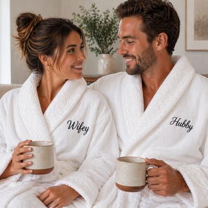 May include: A couple wearing white terry cloth robes embroidered with the words "Wifey" and "Hubby". They are holding beige mugs with brown bases. The robes have shawl collars and are made of a soft, textured material, perfect for relaxing.