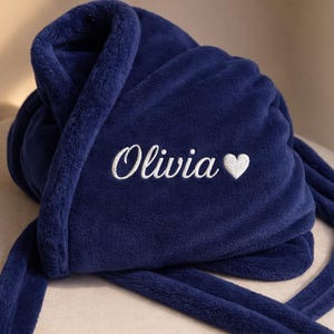 May include: A navy blue plush robe with the name "Olivia" embroidered in white script, followed by a white heart symbol. The robe is folded, showcasing its soft texture and long tie.