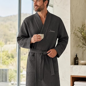 May include: Dark gray waffle-weave robe with a shawl collar and a matching belt. The robe has two front pockets and the name "James" embroidered on the chest. The robe is made of a textured fabric.