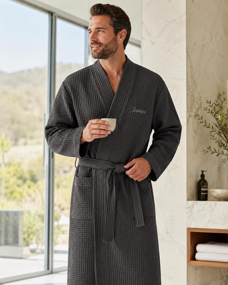 May include: Dark gray waffle-weave robe with a shawl collar and a matching belt. The robe has two front pockets and the name "James" embroidered on the chest. The robe is made of a textured fabric.