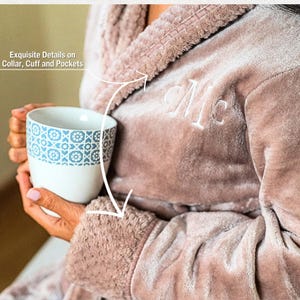 May include: A plush, mauve-colored robe with embroidered initials and a cozy collar and cuffs. The robe is held by a person, who is also holding a white mug with a blue geometric pattern. The text "Exquisite Details on Collar, Cuff and Pockets" is visible.