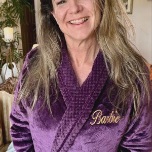 May include: A plush, purple robe with a shawl collar. The robe features the name "Barbie" embroidered in gold script on the chest. The robe is made of a soft, velvety material.