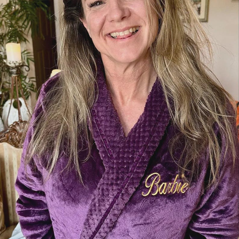 May include: A plush, purple robe with a shawl collar. The robe features the name "Barbie" embroidered in gold script on the chest. The robe is made of a soft, velvety material.