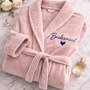 May include: A soft, pink plush robe with the word "Bridesmaid" embroidered in blue script, along with a blue heart. The robe has a shawl collar, a tie belt, and textured cuffs. A bottle of perfume and white roses are also in the image.