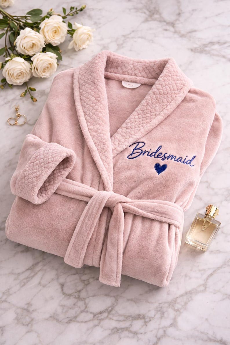 May include: A soft, pink plush robe with the word "Bridesmaid" embroidered in blue script, along with a blue heart. The robe has a shawl collar, a tie belt, and textured cuffs. A bottle of perfume and white roses are also in the image.