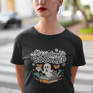 Op de afbeelding: Zwart T-shirt met een geest die een boek leest en de tekst "My weekend is BOOOKED".