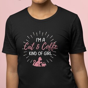 Op de afbeelding: Zwart T-shirt met een roze afbeelding waarop staat "I'm a Cat & Coffee Kind of Girl" met een roze illustratie van een kat en een kopje koffie.