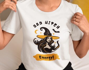 Può includere: Una t-shirt bianca a maniche corte con una stampa grafica di una strega che indossa un cappello a tesa larga. Il testo "BAD WITCH" è scritto sopra l'illustrazione e "Energy!" su uno sfondo a pennellata gialla sotto, con una luna e un pipistrello.
