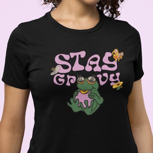 Puede incluir: Camiseta negra con un gráfico rosa "Stay Groovy" que presenta una rana verde con gafas de sol y una flor rosa. El gráfico también incluye una mariposa y una abeja.
