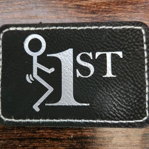 May include: Black rectangular patch with a white stitched border. The patch features a silver graphic of a stick figure and the number "1ST". The patch is made of a textured material.
