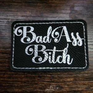 May include: Black rectangular patch with the words "Bad Ass Bitch" in white script lettering. The patch has a white stitched border and is set against a dark wood grain background.